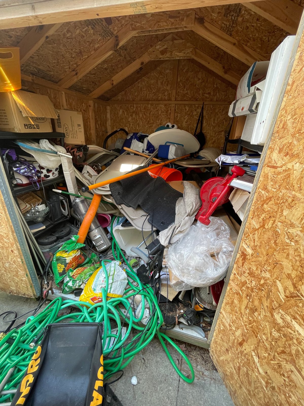 Cluttered wooden shed interior filled with miscellaneous items including an orange bicycle, tangled green cords, tools, and storage boxes
